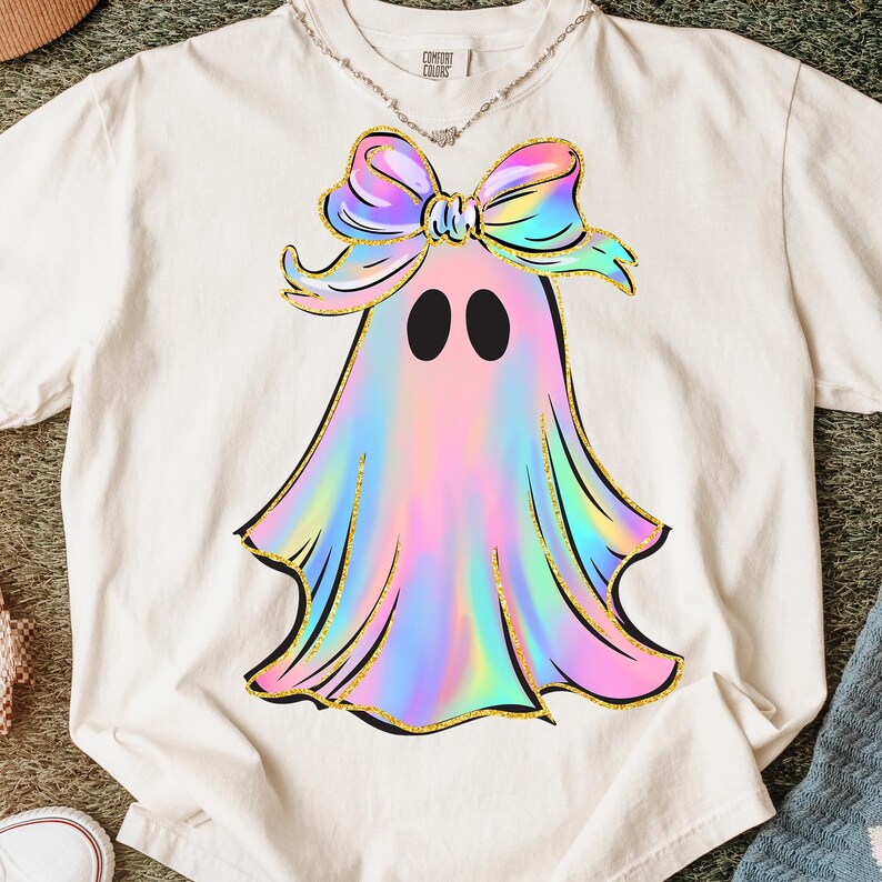 May include: Cream-colored t-shirt featuring a ghost design with a holographic, pastel color scheme. The ghost has a large bow and gold glitter accents. The shirt has the text "Comfort Colors" on the upper back.