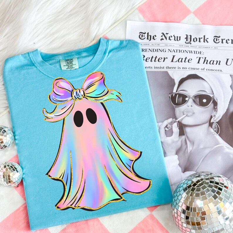 May include: A light blue t-shirt featuring a colorful ghost design with a bow. The ghost has a rainbow-colored body with gold glitter accents. The shirt is on a pink and white checkered surface with a newspaper and disco balls.