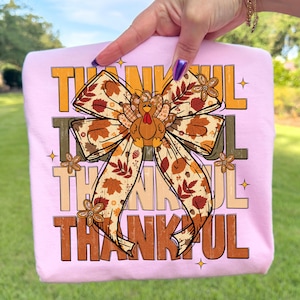 May include: A light pink t-shirt featuring the word "THANKFUL" in a mix of brown and orange block letters. A large bow with a turkey in the center is the focal point. The bow has a fall leaf pattern.
