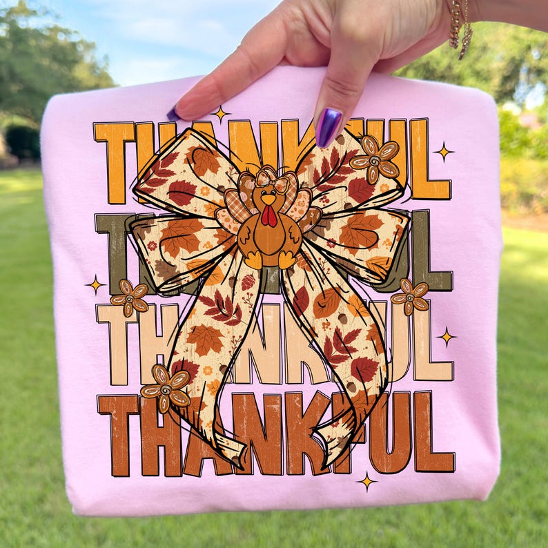 May include: A light pink t-shirt featuring the word "THANKFUL" in a mix of brown and orange block letters. A large bow with a turkey in the center is the focal point. The bow has a fall leaf pattern.