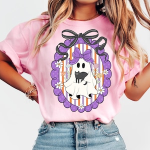 May include: Pink t-shirt featuring a Halloween-themed graphic. The graphic includes a white ghost with a black cat, a purple bow, and a decorative frame with flowers. The background has orange and white stripes.