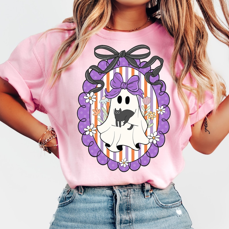 May include: Pink t-shirt featuring a Halloween-themed graphic. The graphic includes a white ghost with a black cat, a purple bow, and a decorative frame with flowers. The background has orange and white stripes.