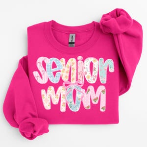 Puede incluir: Una sudadera rosa brillante con las palabras "Senior Mom" en un dise&ntilde;o de patchwork. Las letras est&aacute;n decoradas con telas florales y estampadas, y un gr&aacute;fico de zapatilla de ballet est&aacute; en el centro. La sudadera tiene cuello redondo y mangas largas.