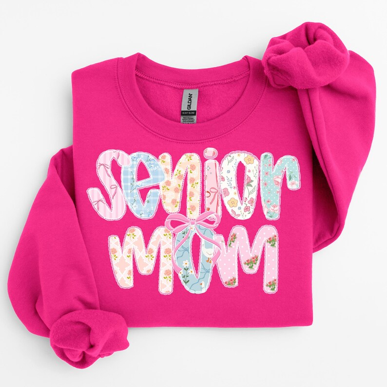 Puede incluir: Una sudadera rosa brillante con las palabras "Senior Mom" en un dise&ntilde;o de patchwork. Las letras est&aacute;n decoradas con telas florales y estampadas, y un gr&aacute;fico de zapatilla de ballet est&aacute; en el centro. La sudadera tiene cuello redondo y mangas largas.