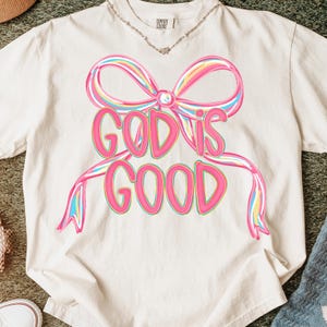 May include: Cream-colored t-shirt featuring a large, colorful bow design with the words "GOD IS GOOD" in a pink, outlined font. The bow has pink, yellow, and blue accents. The shirt is made of a soft material.