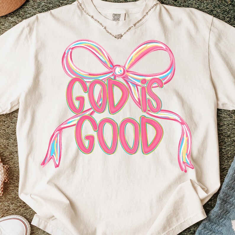 May include: Cream-colored t-shirt featuring a large, colorful bow design with the words "GOD IS GOOD" in a pink, outlined font. The bow has pink, yellow, and blue accents. The shirt is made of a soft material.