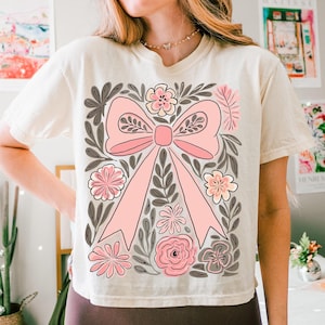 K&ouml;nnte beinhalten: Cremefarbenes T-Shirt mit einem gro&szlig;en rosa Schleifendesign, umgeben von floralen und Blattillustrationen in Rosa, Grau und Creme. Das kurz&auml;rmelige Shirt hat einen Rundhalsausschnitt.