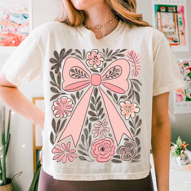 K&ouml;nnte beinhalten: Cremefarbenes T-Shirt mit einem gro&szlig;en rosa Schleifendesign, umgeben von floralen und Blattillustrationen in Rosa, Grau und Creme. Das kurz&auml;rmelige Shirt hat einen Rundhalsausschnitt.