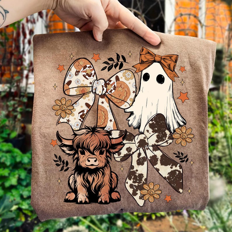 Op de afbeelding: Een bruin t-shirt met een grillig ontwerp met een spook, een Schotse hooglander en decoratieve strikken. Het ontwerp bevat bloemen- en steraccenten, met een kleurenpalet van bruin, oranje en cr&egrave;me.