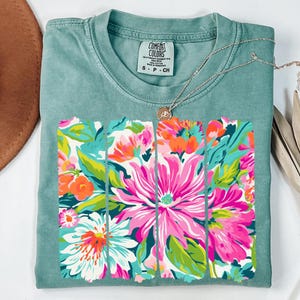 May include: A teal blue t-shirt with a floral print design. The print features a white background with pink, orange, and green flowers. The shirt has a crew neck and short sleeves. The text "COMFORT COLORS" is visible on the tag.