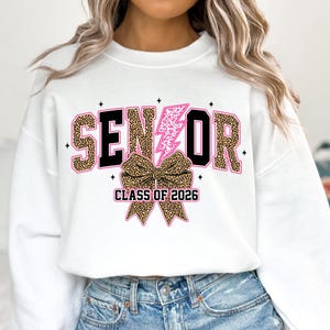Pu&ograve; includere: Felpa bianca con la scritta "SENIOR" in un font leopardato, bordata di nero, con un fulmine rosa e un fiocco leopardato. Il testo "CLASS OF 2026" &egrave; sotto il fiocco.
