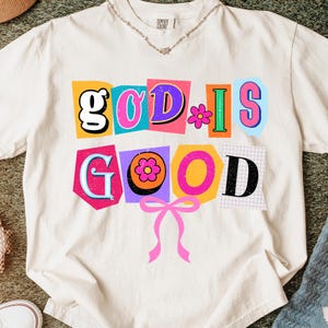 May include: Cream-colored t-shirt with the phrase "GOD IS GOOD" in colorful, retro-style lettering. The design features a pink bow at the bottom and a small flower graphic. The shirt has a relaxed fit.