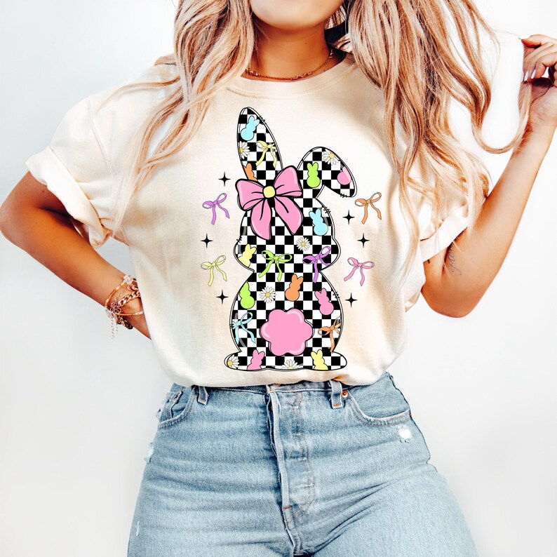 May include: A white t-shirt with a checkered bunny silhouette design. The bunny is decorated with pastel-colored bows, flowers, and candy.