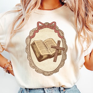May include: Cream-colored t-shirt featuring a framed illustration of an open book and a cross. The illustration is in a vintage style, with a pink bow at the top and a decorative border. The design has a religious theme.