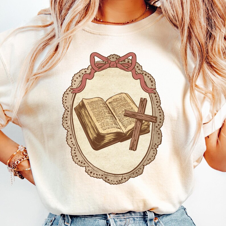 May include: Cream-colored t-shirt featuring a framed illustration of an open book and a cross. The illustration is in a vintage style, with a pink bow at the top and a decorative border. The design has a religious theme.