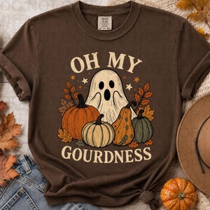 May include: A dark brown short-sleeve t-shirt featuring a graphic of a white ghost surrounded by orange, yellow, and green pumpkins and autumn leaves. The text reads OH MY GOURDNESS in a cream-colored font above and below the central illustration.