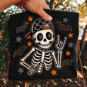 May include: A black t-shirt featuring a graphic of a skeleton wearing a black and gray checkered baseball cap. The skeleton is making a rock and roll hand gesture. The design includes orange and gray stars and black bats on a black background.