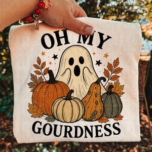 May include: A cream-colored t-shirt featuring a graphic print of a ghost, three pumpkins, and autumn leaves. The text reads OH MY GOURDNESS in black lettering. The design includes small black and orange stars surrounding the central ghost figure.