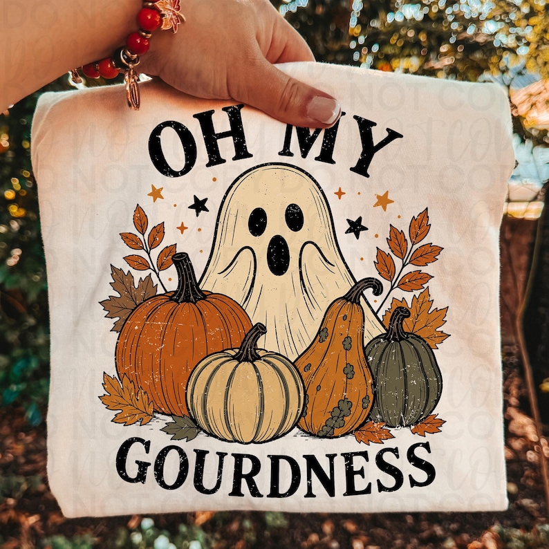 May include: A cream-colored t-shirt featuring a graphic print of a ghost, three pumpkins, and autumn leaves. The text reads OH MY GOURDNESS in black lettering. The design includes small black and orange stars surrounding the central ghost figure.