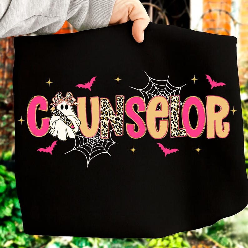 Pu&ograve; includere: Borsa tote nera con la scritta "COUNSELOR" in lettere rosa e beige. La lettera "U" presenta un motivo leopardato. Un fantasma dei cartoni animati con un fiocco e una matita &egrave; a sinistra. Pipistrelli rosa, ragnatele e stelle decorano il disegno.