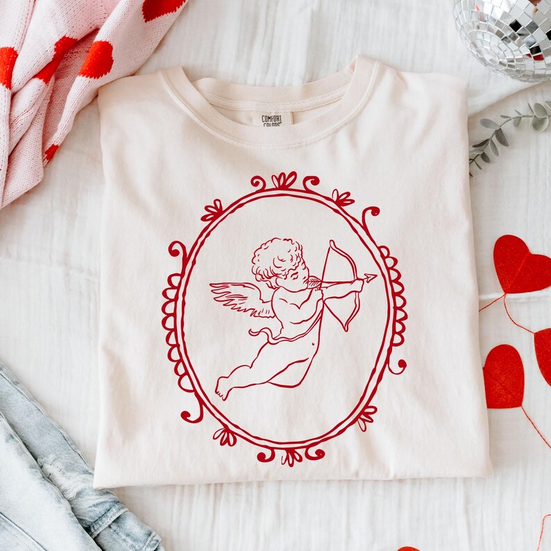 Pu&ograve; includere: T-shirt color crema con un'illustrazione rossa di Cupido che punta il suo arco e le frecce all'interno di una cornice ovale. La maglietta &egrave; piegata su una superficie bianca, con decorazioni di San Valentino sullo sfondo.