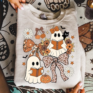 Op de afbeelding: Beige sweatshirt met een Halloween-thema. Het ontwerp bevat twee stripgeesten die boeken lezen, omringd door strikken, bloemen, pompoenen, sterren en een zwarte kat. De strikken hebben een luipaardprint.