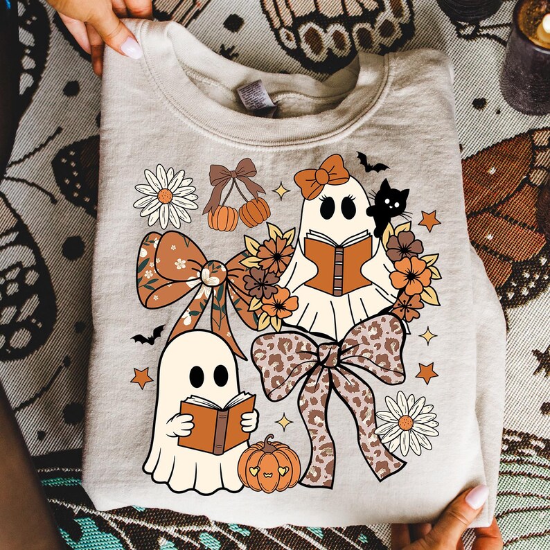 Op de afbeelding: Beige sweatshirt met een Halloween-thema. Het ontwerp bevat twee stripgeesten die boeken lezen, omringd door strikken, bloemen, pompoenen, sterren en een zwarte kat. De strikken hebben een luipaardprint.