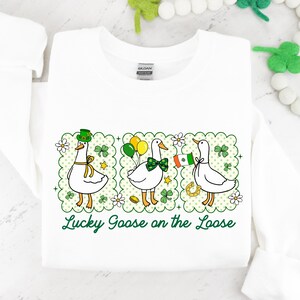 Peut inclure: Sweat-shirt blanc avec un motif de la Saint-Patrick. Trois oies de dessin anim&eacute; sont repr&eacute;sent&eacute;es, chacune avec des accessoires festifs : un chapeau vert, des ballons et un drapeau. Le texte "Lucky Goose on the Loose" est &eacute;crit en vert.