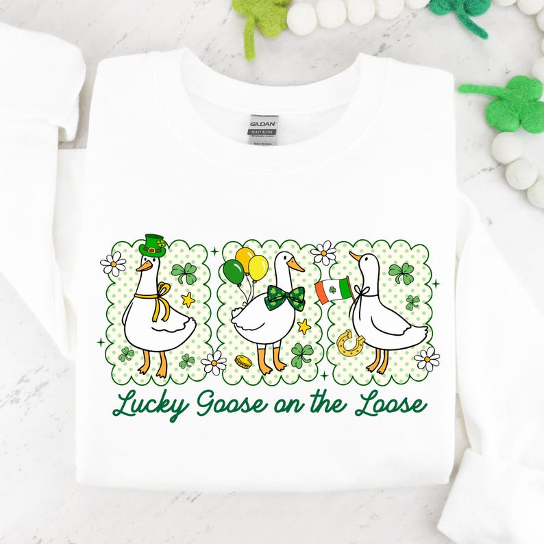 Peut inclure: Sweat-shirt blanc avec un motif de la Saint-Patrick. Trois oies de dessin anim&eacute; sont repr&eacute;sent&eacute;es, chacune avec des accessoires festifs : un chapeau vert, des ballons et un drapeau. Le texte "Lucky Goose on the Loose" est &eacute;crit en vert.