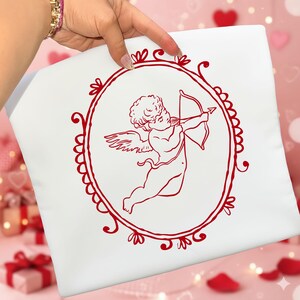 Pu&ograve; includere: Cuscino bianco con un'illustrazione rossa di Cupido che scocca una freccia con l'arco. L'immagine di Cupido &egrave; racchiusa in una cornice ovale rossa con decorazioni. Il cuscino &egrave; tenuto da una persona.