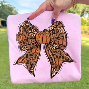 May include: A light pink sweatshirt featuring a large bow design. The bow has a leopard print pattern with orange pumpkins and white flowers. The bow is outlined in black. The sweatshirt is held up by a hand.