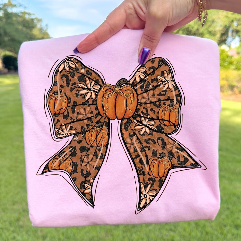 May include: A light pink sweatshirt featuring a large bow design. The bow has a leopard print pattern with orange pumpkins and white flowers. The bow is outlined in black. The sweatshirt is held up by a hand.