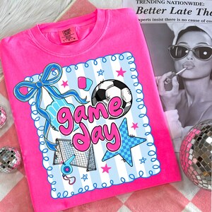 May include: A bright pink Comfort Colors sweatshirt featuring a soccer-themed graphic. The graphic includes a soccer ball, goal, and the words "Game Day" in a playful font, with blue and pink accents. The shirt is a crew neck.
