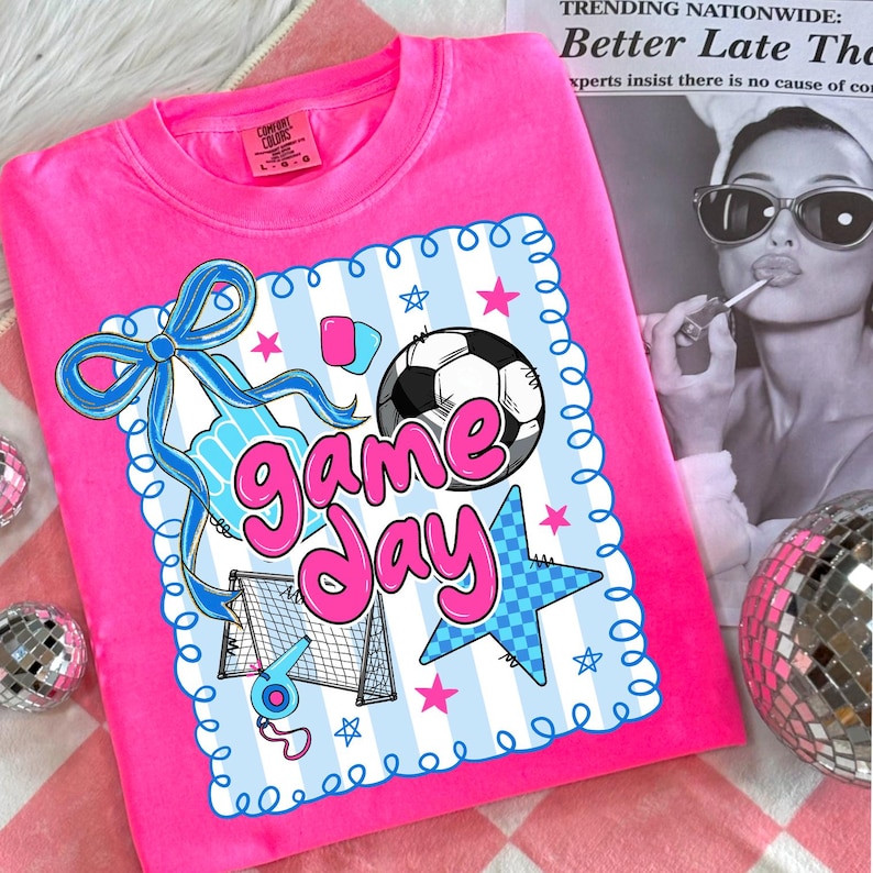 May include: A bright pink Comfort Colors sweatshirt featuring a soccer-themed graphic. The graphic includes a soccer ball, goal, and the words "Game Day" in a playful font, with blue and pink accents. The shirt is a crew neck.