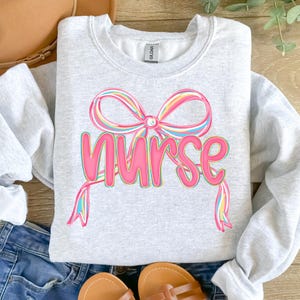 Pode incluir: Camisola cinza claro com a palavra "Nurse" em rosa, com um design de la&ccedil;o colorido por cima. O la&ccedil;o tem listras coloridas do arco-&iacute;ris. A camisola &eacute; feita de um material macio, ideal para profissionais de sa&uacute;de.