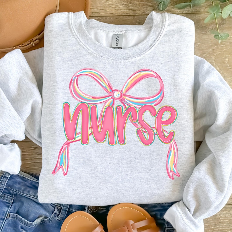 Pode incluir: Camisola cinza claro com a palavra "Nurse" em rosa, com um design de la&ccedil;o colorido por cima. O la&ccedil;o tem listras coloridas do arco-&iacute;ris. A camisola &eacute; feita de um material macio, ideal para profissionais de sa&uacute;de.