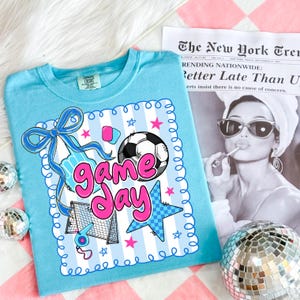 May include: A light blue t-shirt with a soccer-themed graphic that says "Game Day" in pink. The design includes a soccer ball, stars, and a soccer net. The shirt is displayed on a pink and white checkered surface.