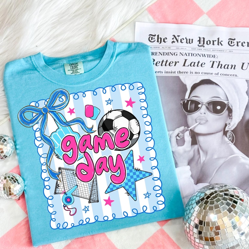 May include: A light blue t-shirt with a soccer-themed graphic that says "Game Day" in pink. The design includes a soccer ball, stars, and a soccer net. The shirt is displayed on a pink and white checkered surface.