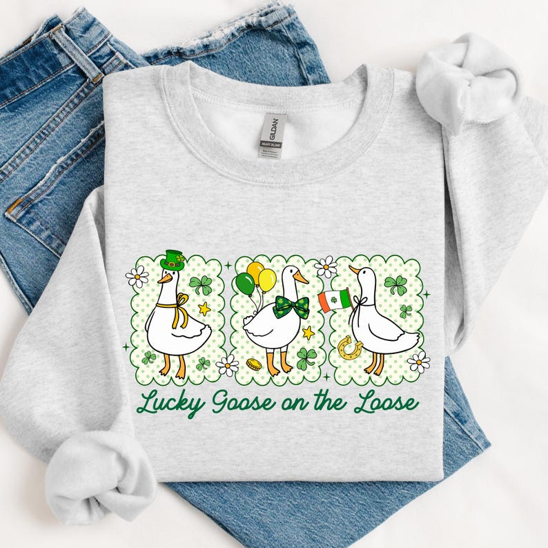 Peut inclure: Un sweat-shirt gris clair avec un motif de trois oies de dessin anim&eacute;, chacune avec un accessoire diff&eacute;rent, et le texte "Lucky Goose on the Loose" en vert. Les oies sont dans une bordure verte festonn&eacute;e avec des tr&egrave;fles et des fleurs.