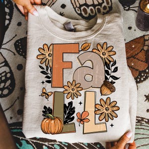 May include: A beige sweatshirt with the word "FALL" in large, stylized letters. The design includes fall-themed graphics like pumpkins, flowers, leaves, butterflies, and acorns in shades of orange, brown, and beige.