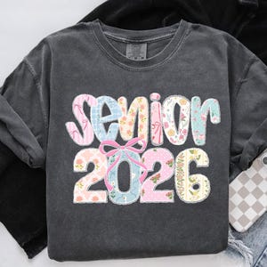 Puede incluir: Sudadera gris oscuro con la palabra "Senior" y el a&ntilde;o "2026" en un dise&ntilde;o floral y estampado colorido. La palabra "Senior" est&aacute; en patrones florales rosas, azules y blancos. El a&ntilde;o "2026" est&aacute; en patrones florales rosas y amarillos.