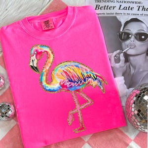 May include: A neon pink sweatshirt featuring a colorful flamingo design with gold glitter accents. The flamingo has a rainbow-colored body and a black beak. The shirt has the Comfort Colors brand tag.