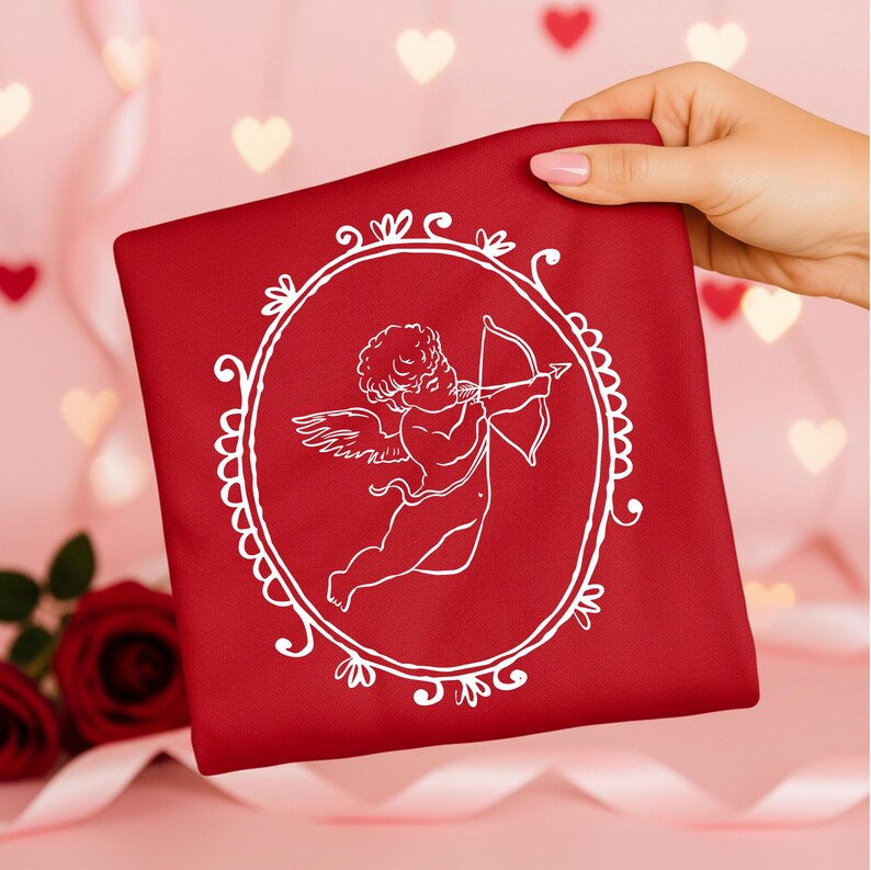 Pu&ograve; includere: Un tessuto quadrato rosso con un'illustrazione bianca di Cupido che scocca una freccia con l'arco, racchiusa da un bordo ovale decorativo. Il disegno &egrave; centrato sul tessuto. Lo sfondo &egrave; rosa tenue con bokeh a forma di cuore.