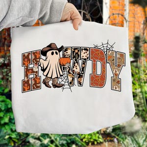 Puede incluir: Sudadera blanca con la palabra "HOWDY" en un dise&ntilde;o de tem&aacute;tica occidental. Los contornos de las letras est&aacute;n rellenos con patrones de cactus, piel de vaca y hojas de oto&ntilde;o. Un fantasma con sombrero y botas de vaquero sostiene una calabaza, con una telara&ntilde;a.