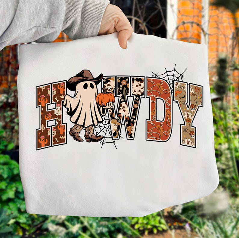 Puede incluir: Sudadera blanca con la palabra "HOWDY" en un dise&ntilde;o de tem&aacute;tica occidental. Los contornos de las letras est&aacute;n rellenos con patrones de cactus, piel de vaca y hojas de oto&ntilde;o. Un fantasma con sombrero y botas de vaquero sostiene una calabaza, con una telara&ntilde;a.