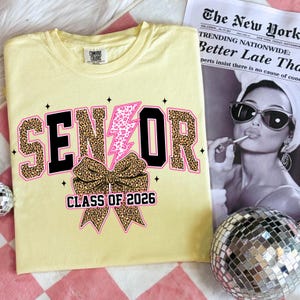 Pu&ograve; includere: Una maglietta giallo chiaro con la scritta "SENIOR" in nero e stampa leopardata, un fulmine rosa e un fiocco leopardato. Il testo "CLASS OF 2026" &egrave; sotto il fiocco. Un giornale e una palla da discoteca sono sullo sfondo.