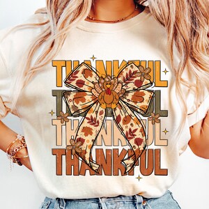 May include: Cream-colored t-shirt with the word "THANKFUL" in layered orange, brown, and tan text. A large bow with fall leaf and turkey designs is centered on the shirt, with small floral accents.