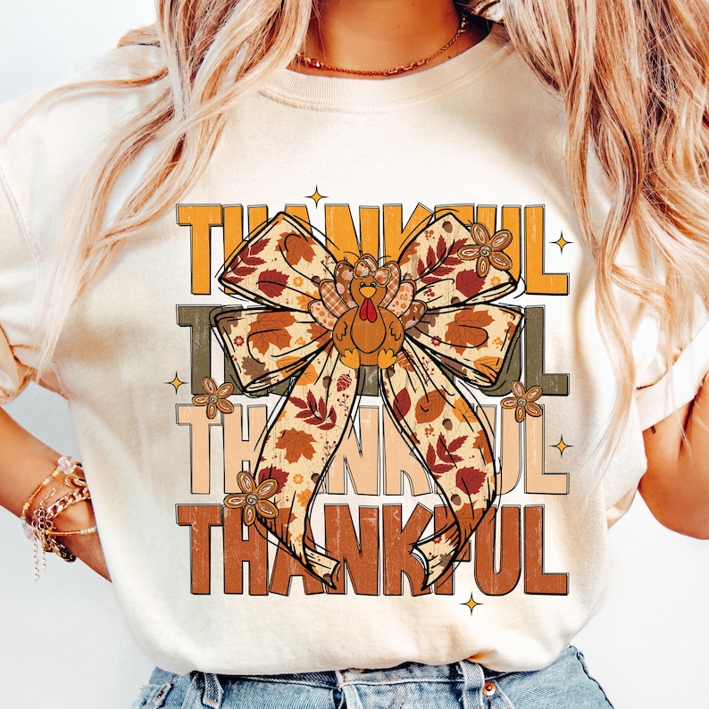 May include: Cream-colored t-shirt with the word "THANKFUL" in layered orange, brown, and tan text. A large bow with fall leaf and turkey designs is centered on the shirt, with small floral accents.
