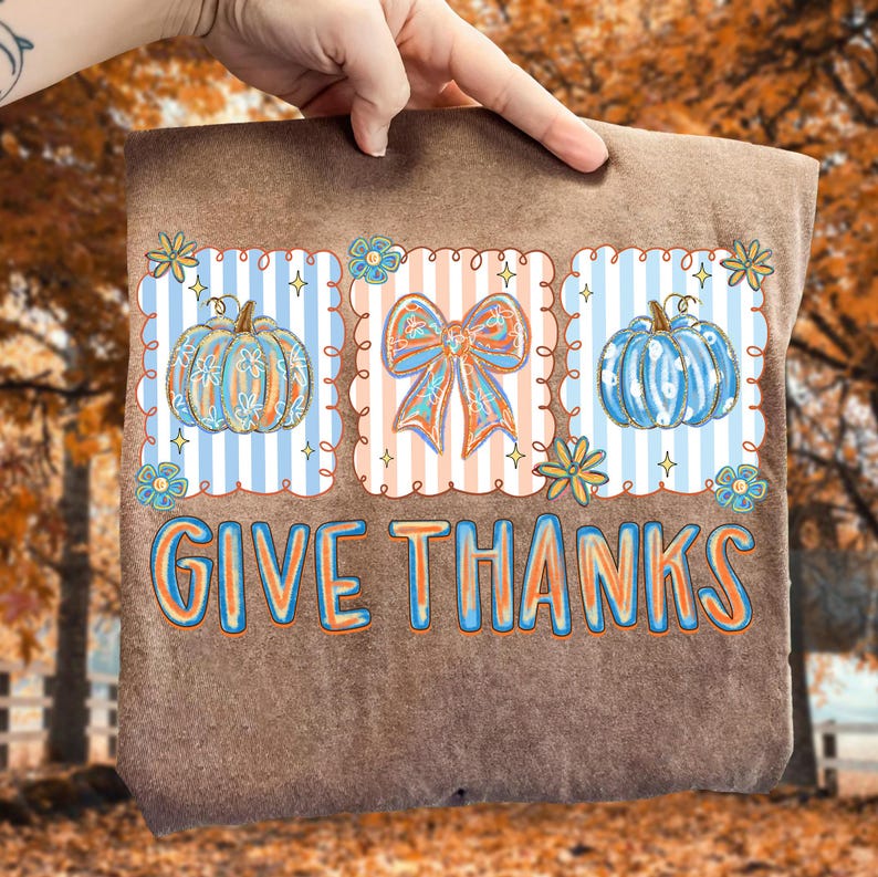 May include: Brown fabric with a colorful graphic that says "GIVE THANKS". The graphic includes pumpkins, a bow, and floral elements. The pumpkins and bow are in orange and blue colors. The background is a fall scene.
