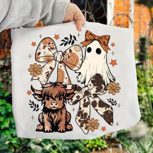 Op de afbeelding: Witte draagtas met een cartoon spook, een Schotse hooglander en twee grote strikken met bloemen- en koeienprintpatronen. Het ontwerp bevat sterren en bloemen in bruin- en oranje tinten.